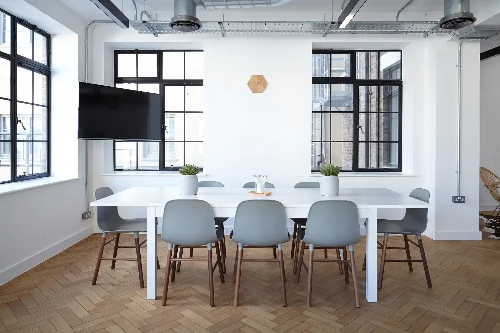 foto van een leeg kantoor. Het is een witte tafel met 6 witte stoelen. In de rechterhoek hangt een beeldscherm.